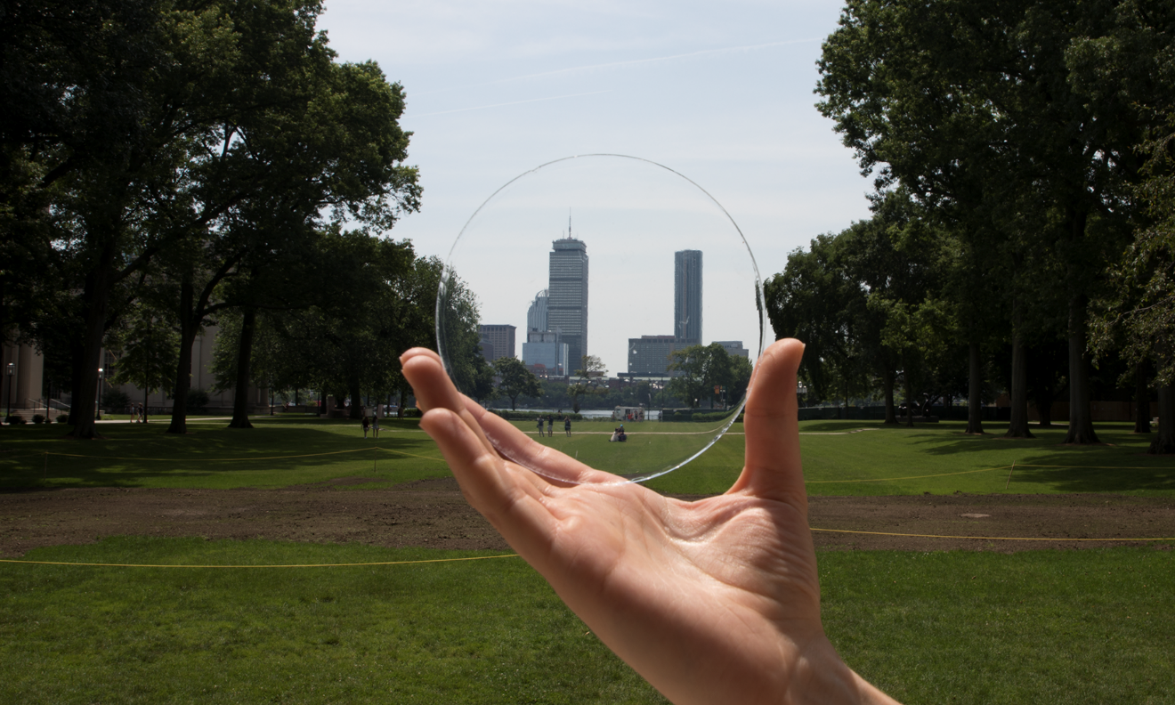 A hand hold a circular piece of Aerigel, which you can see park and city skyline through.