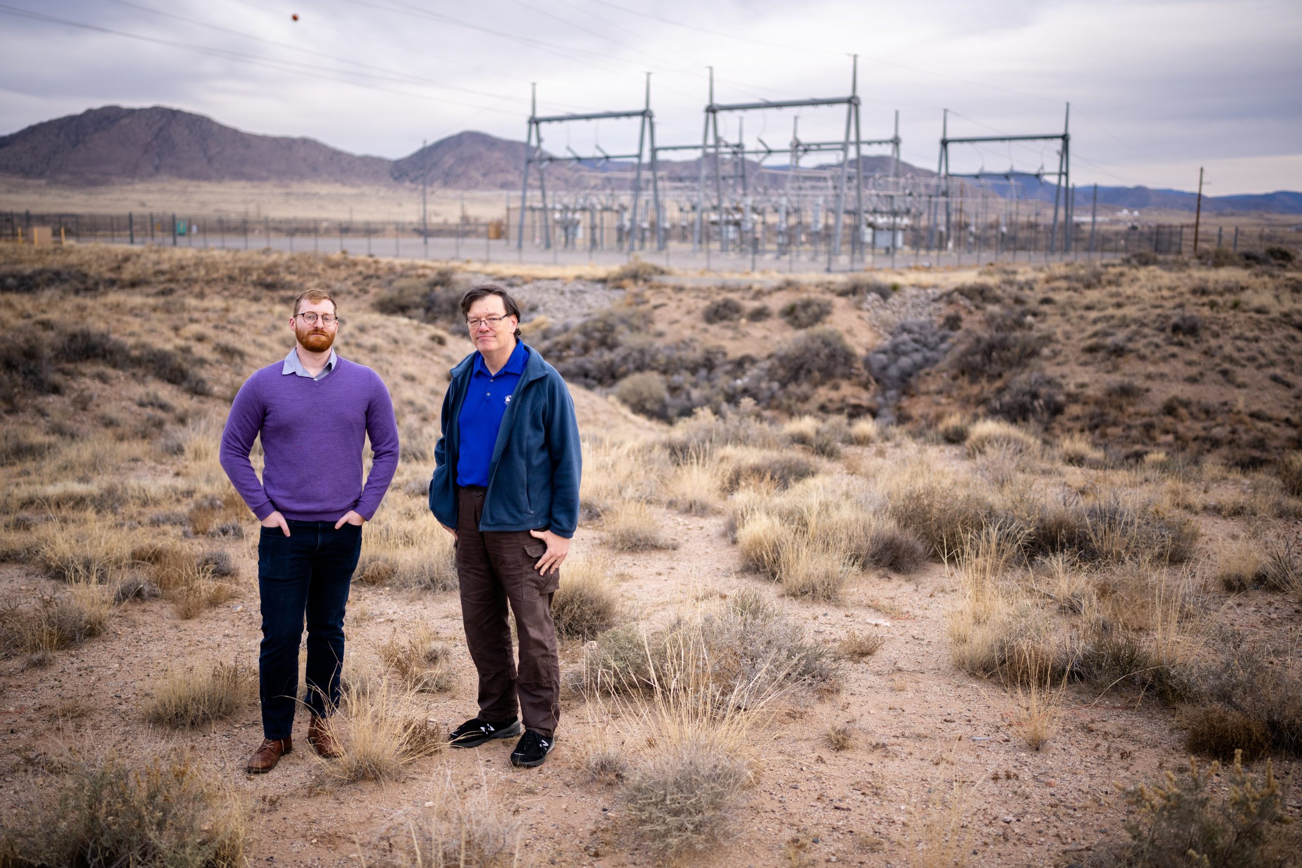 Two researchers stand in the desert. Power transformers are on the horizon in the background.