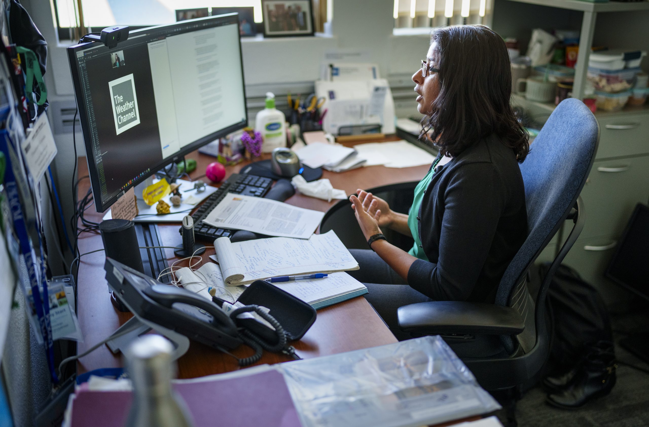 Thushara sits at a desk and uses her computer and webcam to talk to The Weather Channel for a TV segment