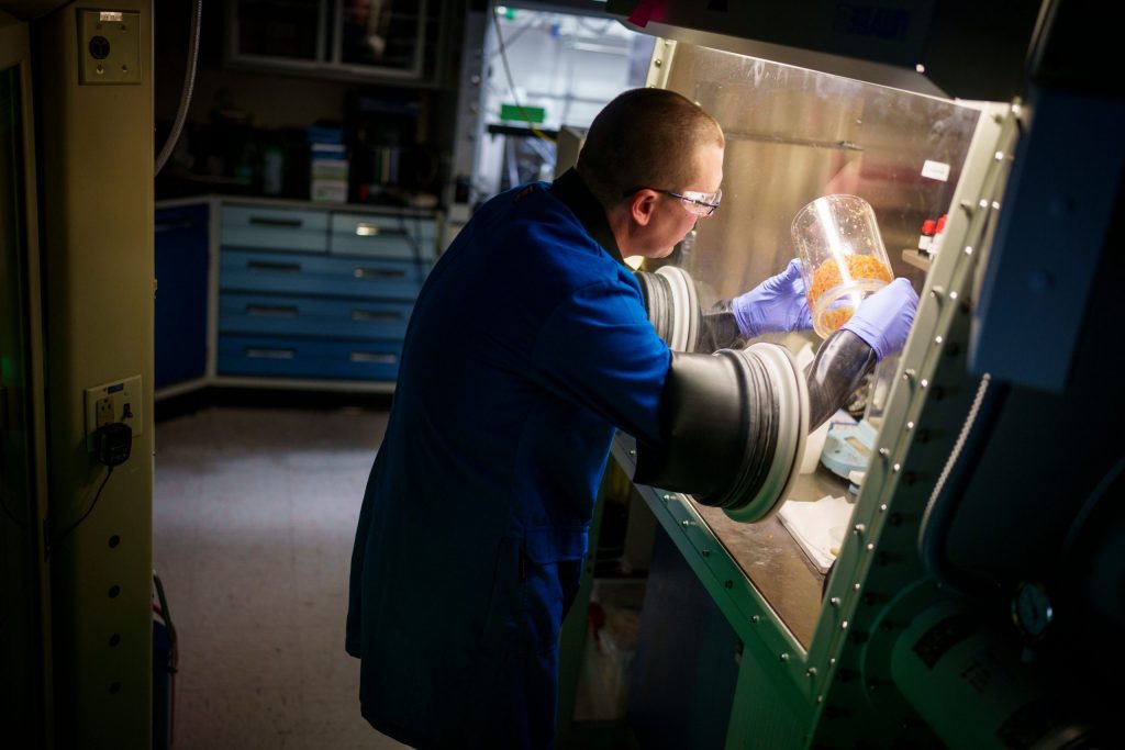 Scientist Cody Corbin stands at a lab station, his body is separated from the material he is working with by glass, and his arms are protected by rubber sleeves.