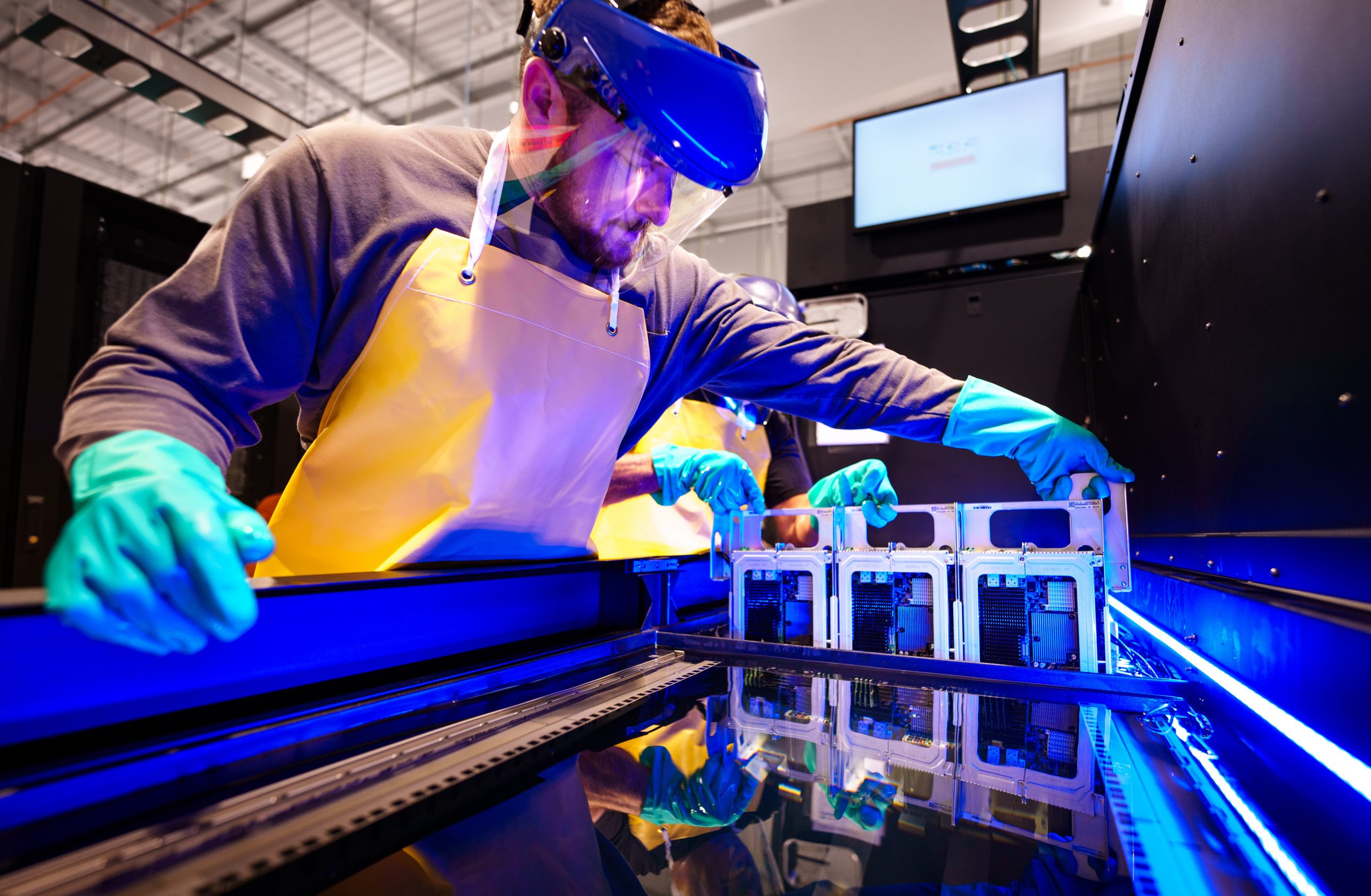 A man with a face shield, apron, and gloves works on a computer server submerged in liquid