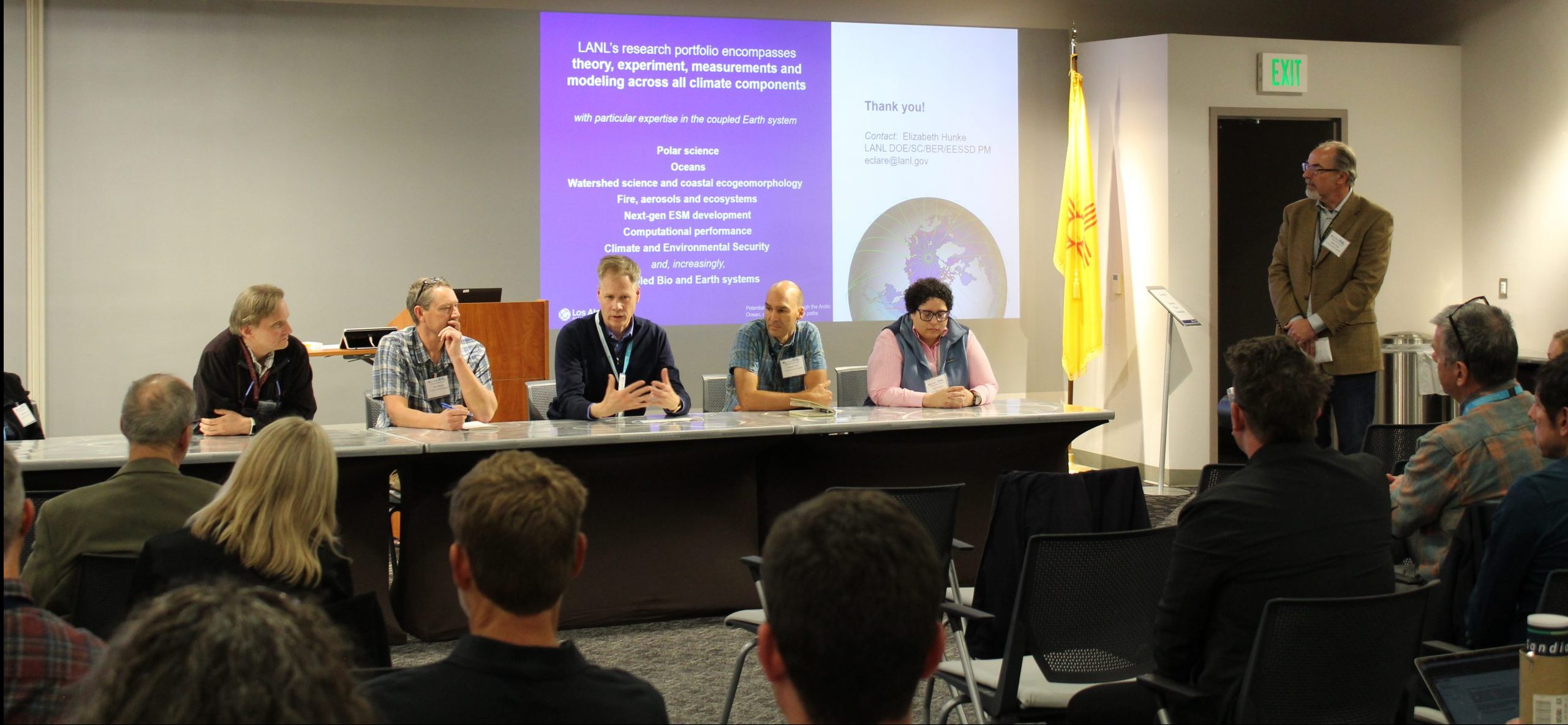 Five scientists sit at a long table in front of a screen and one person stands off to the side with an audience in the foreground