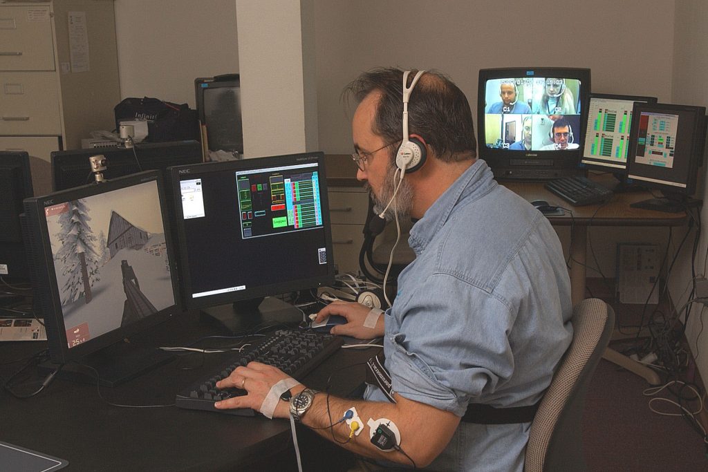 AND YOU’RE FEELING HOW? Sandia researcher Peter Merkle uses sensors to communicate personal biofacts in realtime to a computer as he applies himself to a video game that requires team participation. On split screen to his right are previously imaged photos of Sandia colleagues (clockwise from top left): Simon Goldfine, Adele Doser, Mark Grubelich, and Peter without his beard.