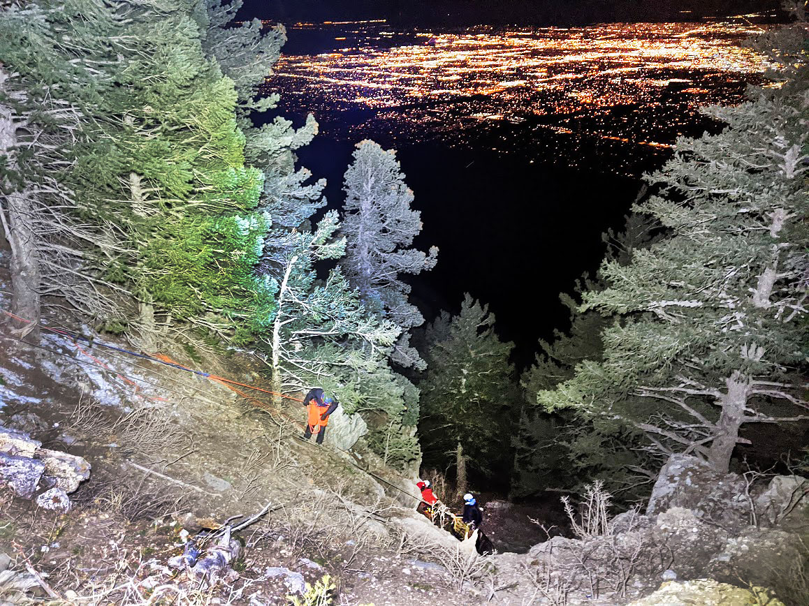 Foregrounds: Pine trees and rocks, with three small figures climbing down a cliffside. Background: the lights of Albuquerque at night.