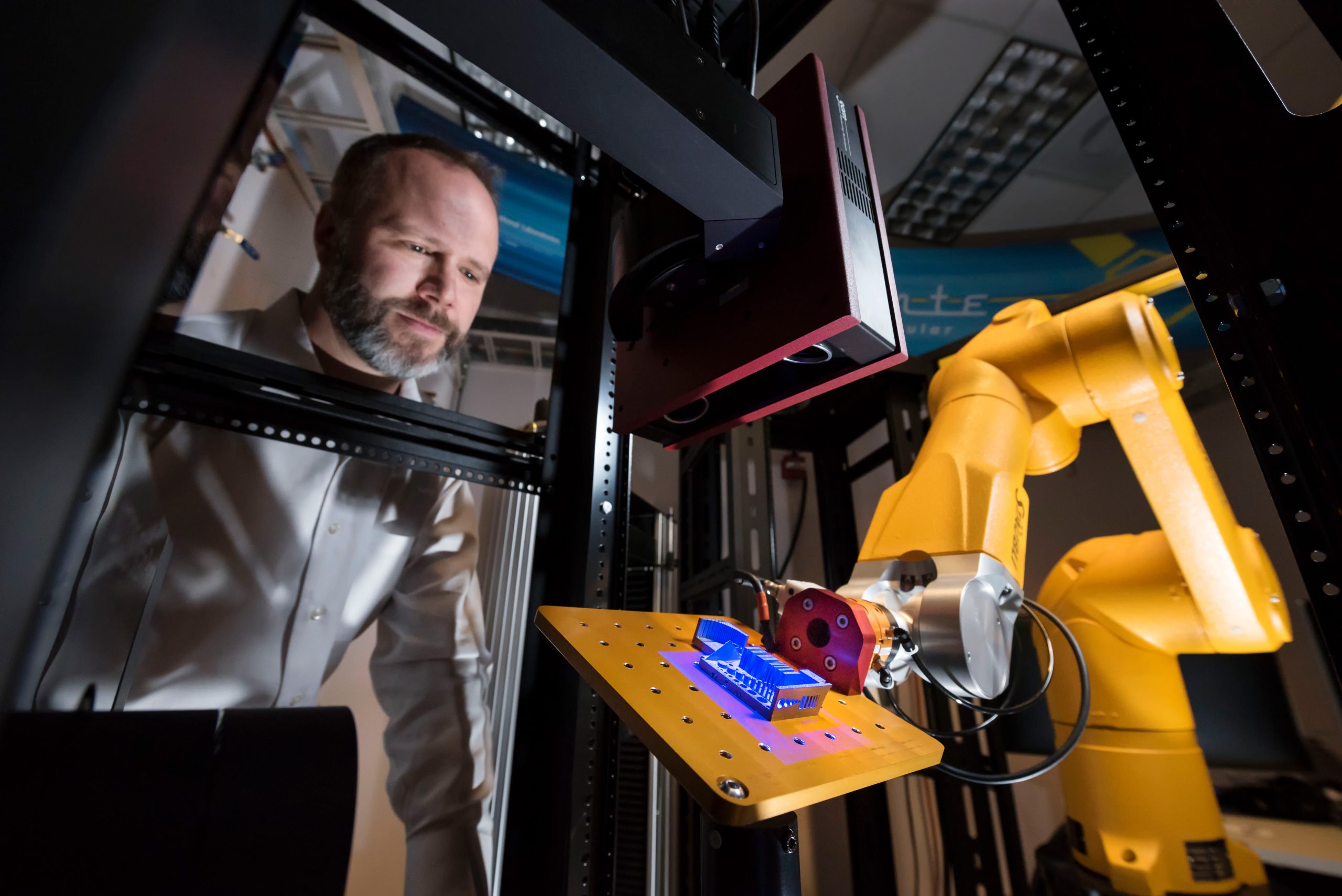 Brad Boyce watches a yellow commercial robot scan a 3D-printed test part with blue light.