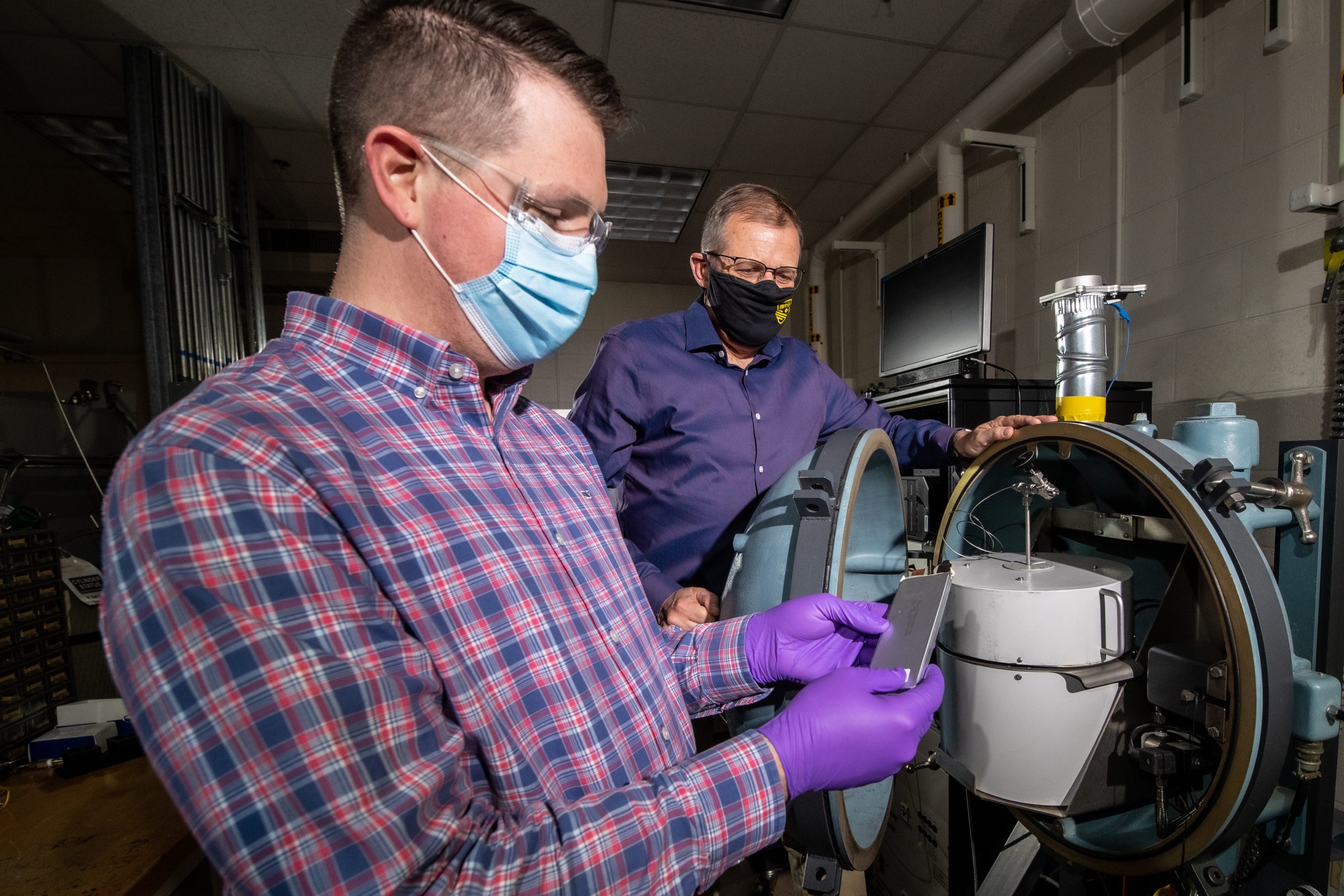 Two men, one holding a shiny battery testing case, stand beside a beach-ball-sized, thick-metal testing instrument.