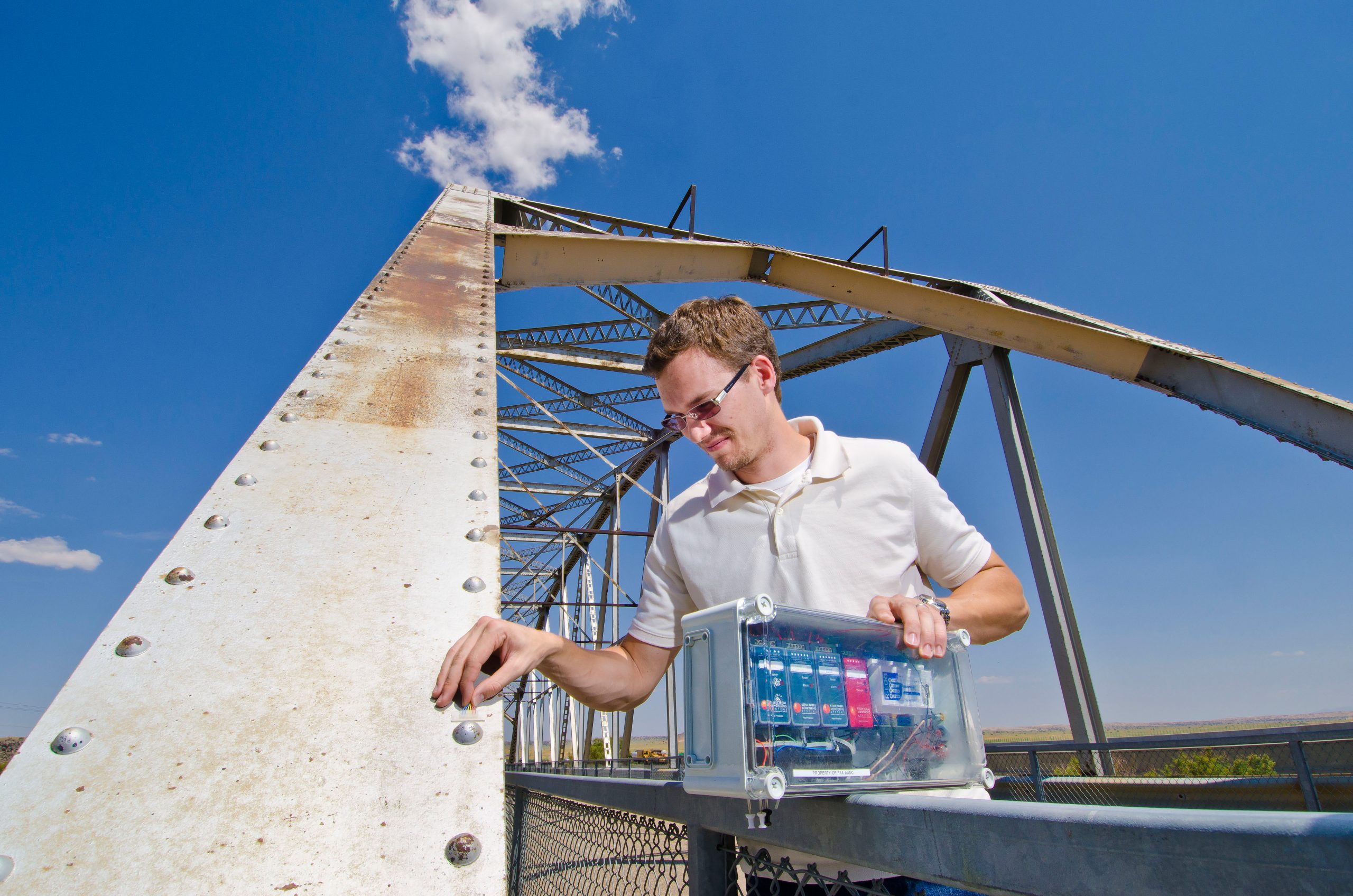 Man positions small, clear sensor on an old rusty bridge. Blue sky in background. In his other hand is a complex control system.