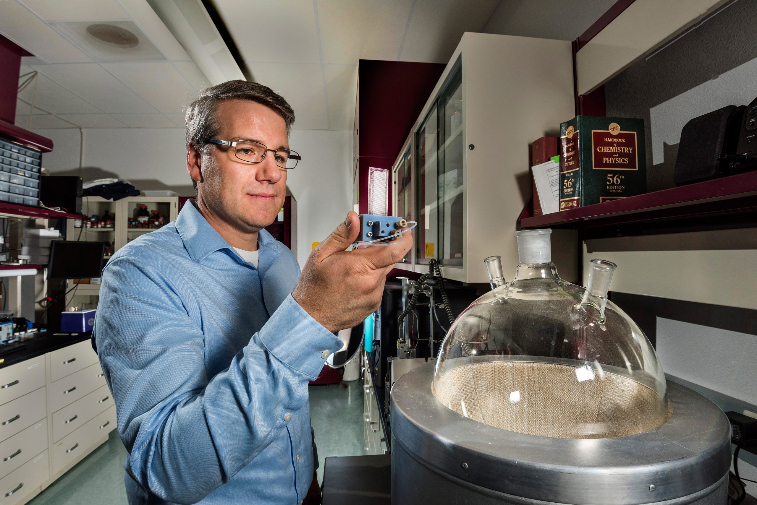 Dale Huber, in a blue labcoat holds a small white microfluidic chip beside a large, basket-ball sized round-bottom flask.