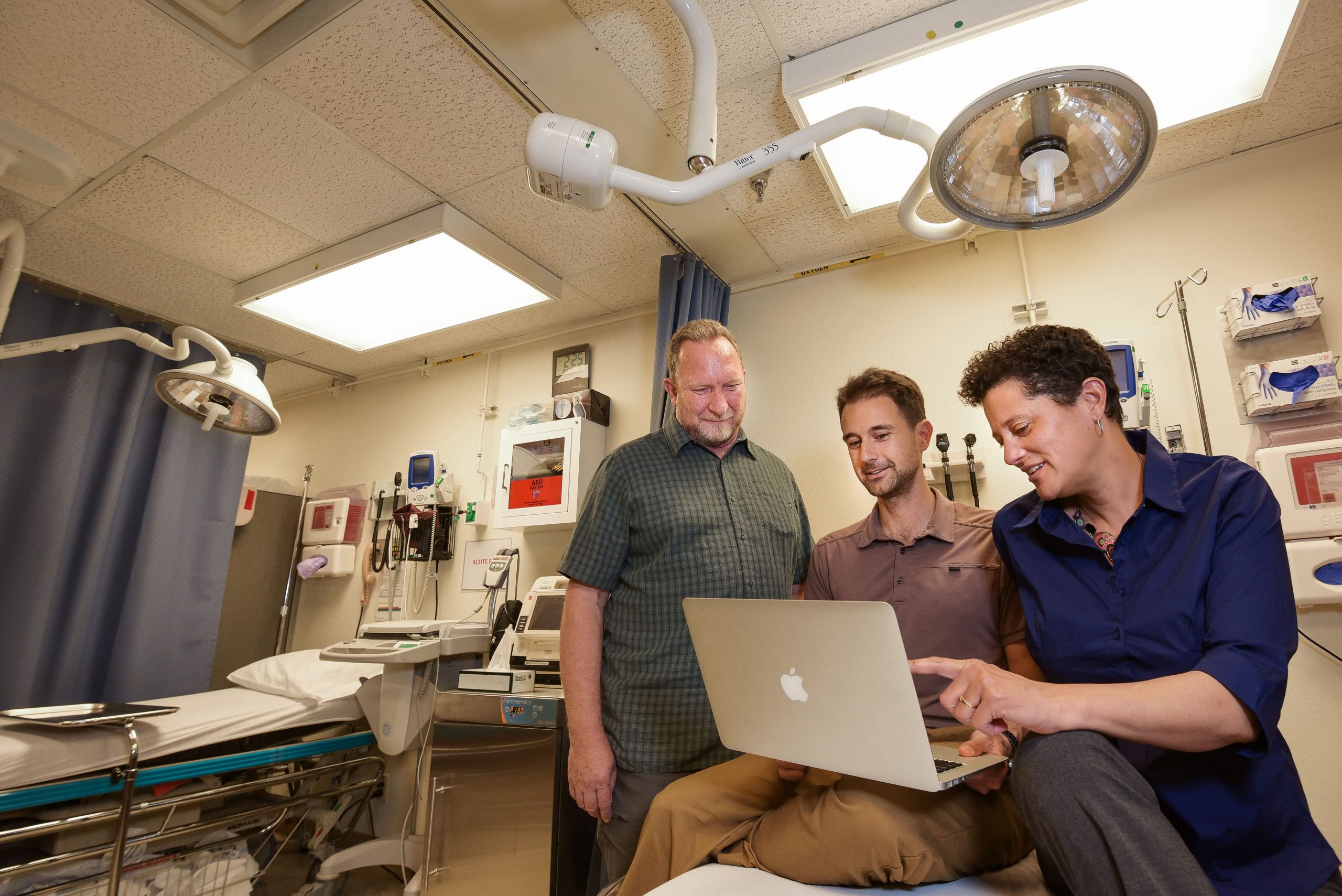 Pat Finley, Drew Levin, and Melanie Moses sit in an emergency room, looking at a laptop.