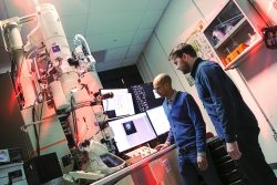 Two men stand near a desk with computer monitors and a towering microscope that reaches the ceiling