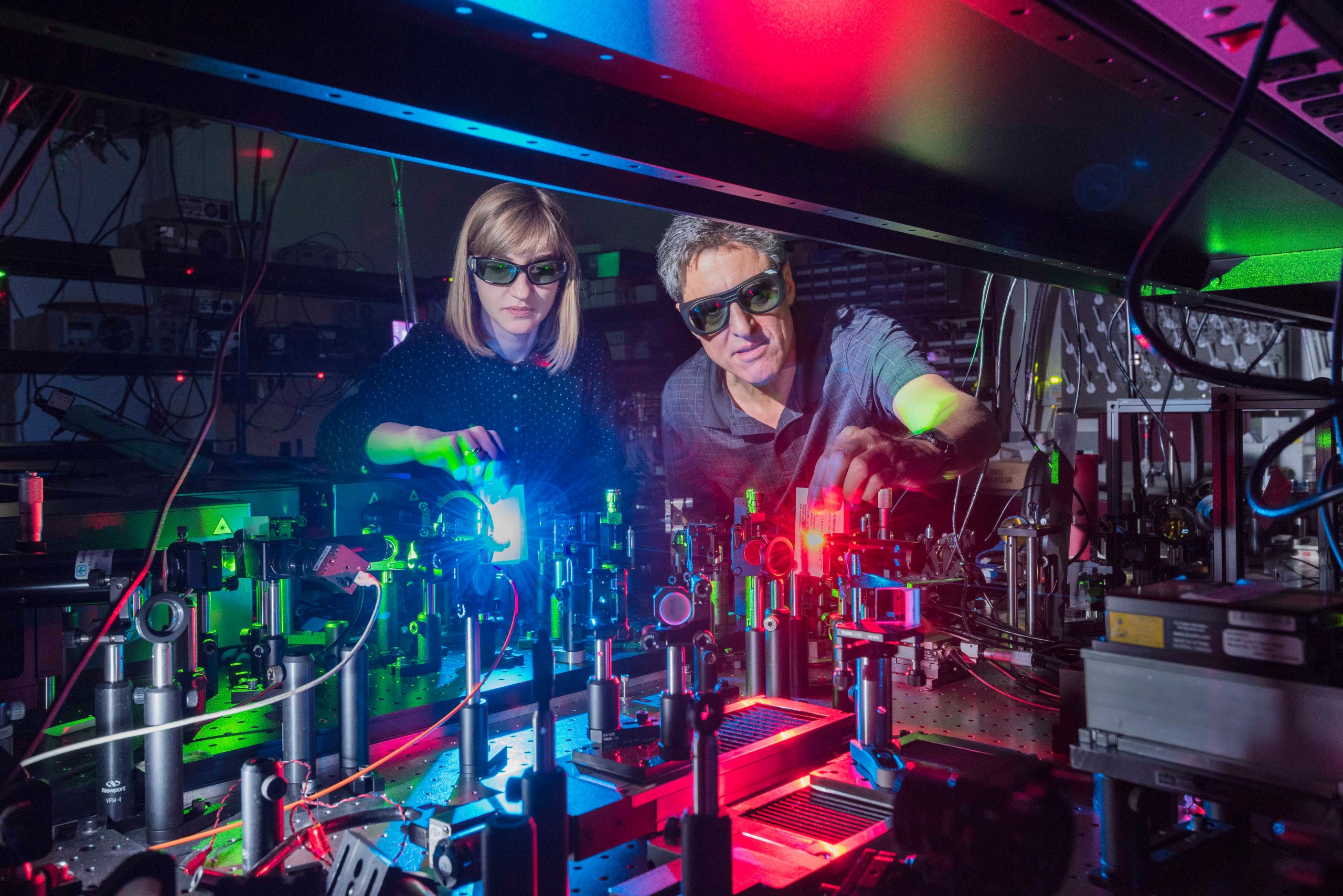 Polina Vabishchevich and Igal Brener in a dark room with blue, green, and red laser light reflecting off of a table full of optical mirrors.