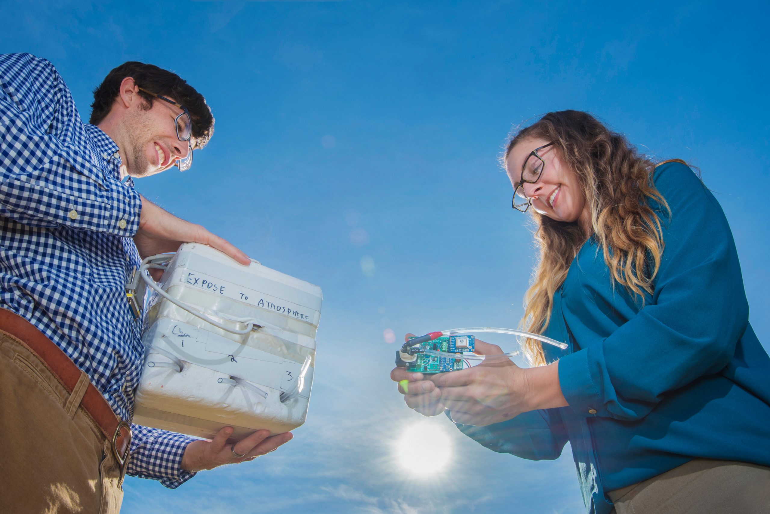 Danny Bowman holding a stryofoam box (left) Sarah Albert holding an infrasound sensor (right) with a bright blue sky as background