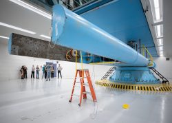 A group of of people stand in the background. In the foreground, a giant blue arm of a mechanical spinning machine sits at rest several feet above the ground.