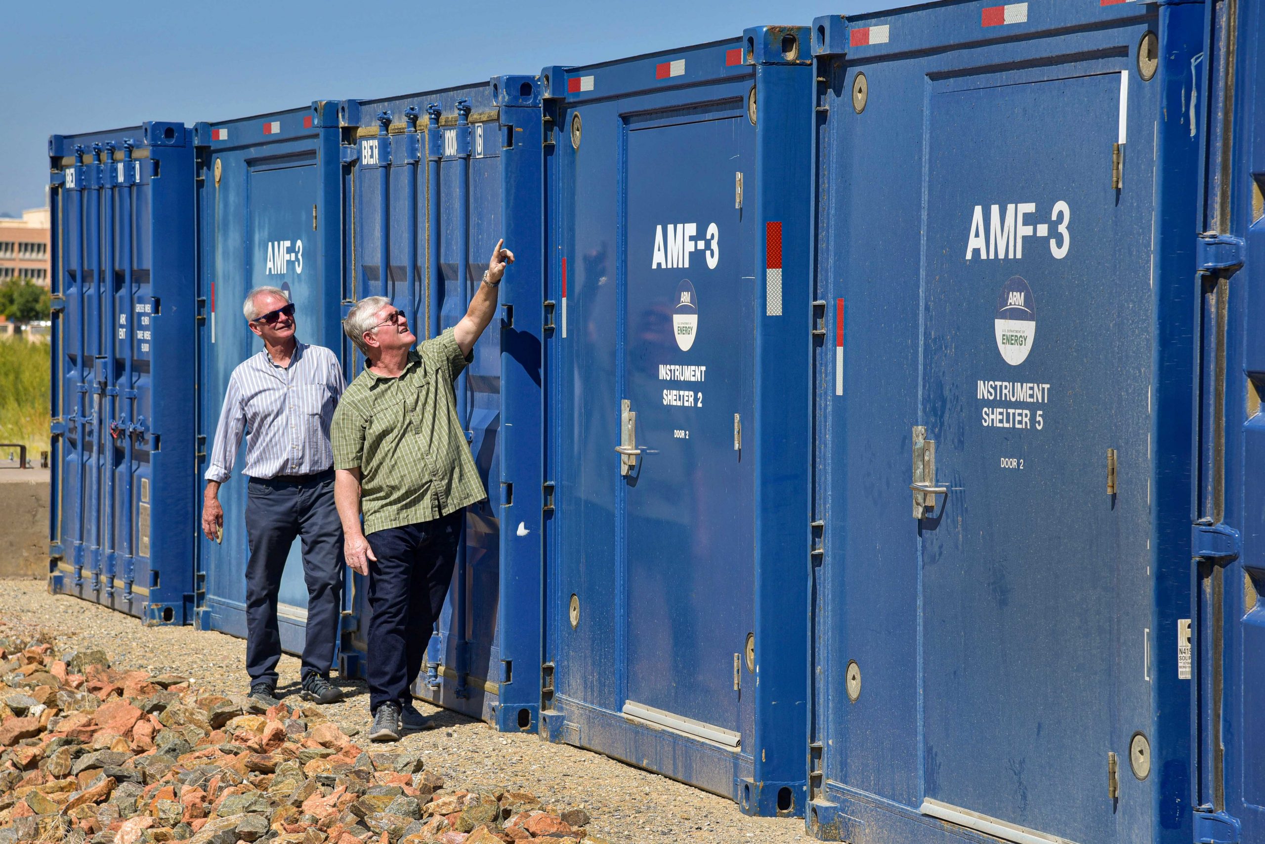 Two men look at blue shipping container-like shelters. One points.