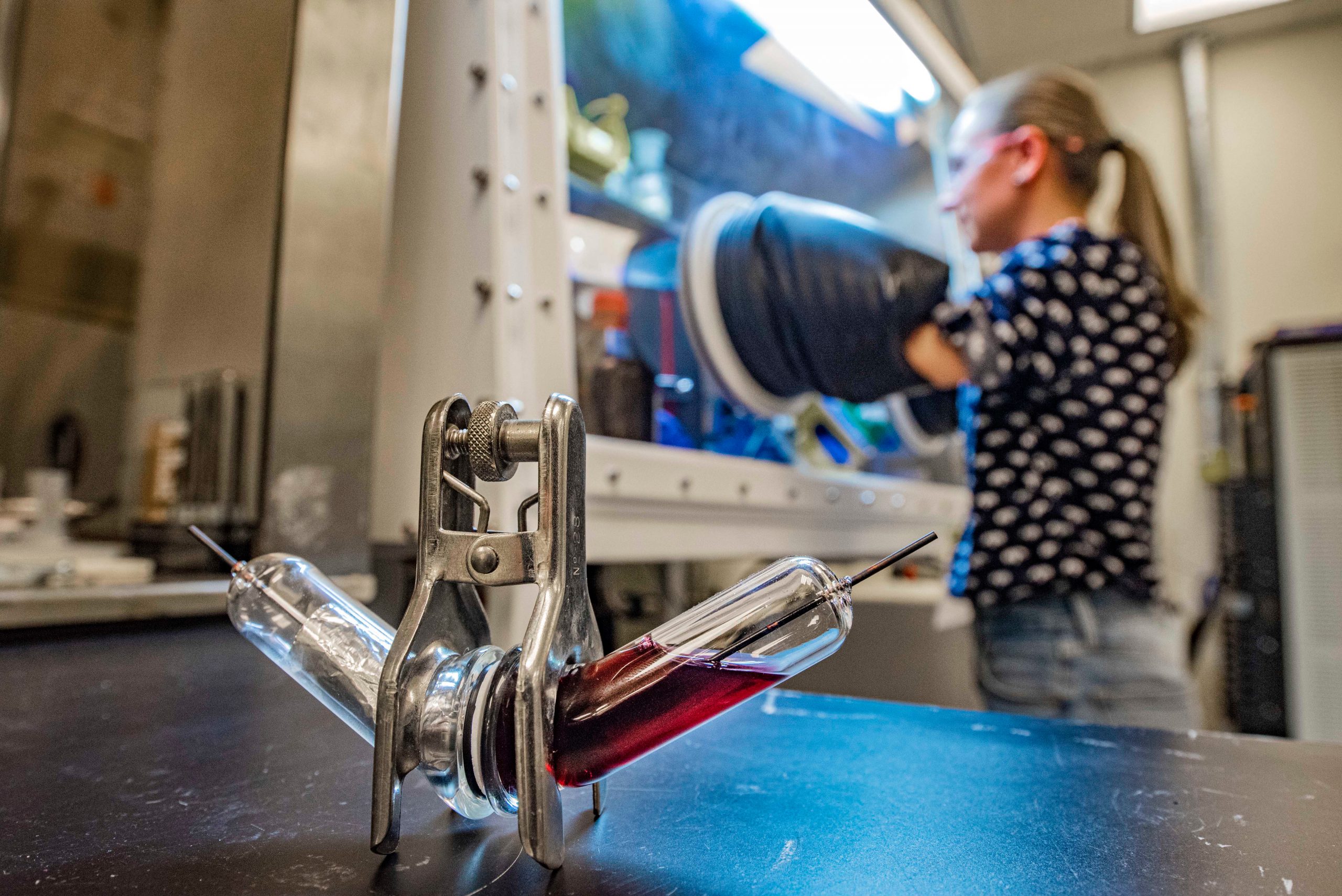 Foreground, two glass tubes connected at an angle with shiny metal in one tube and purple liquid in the other. Background woman with hands in a glove box.