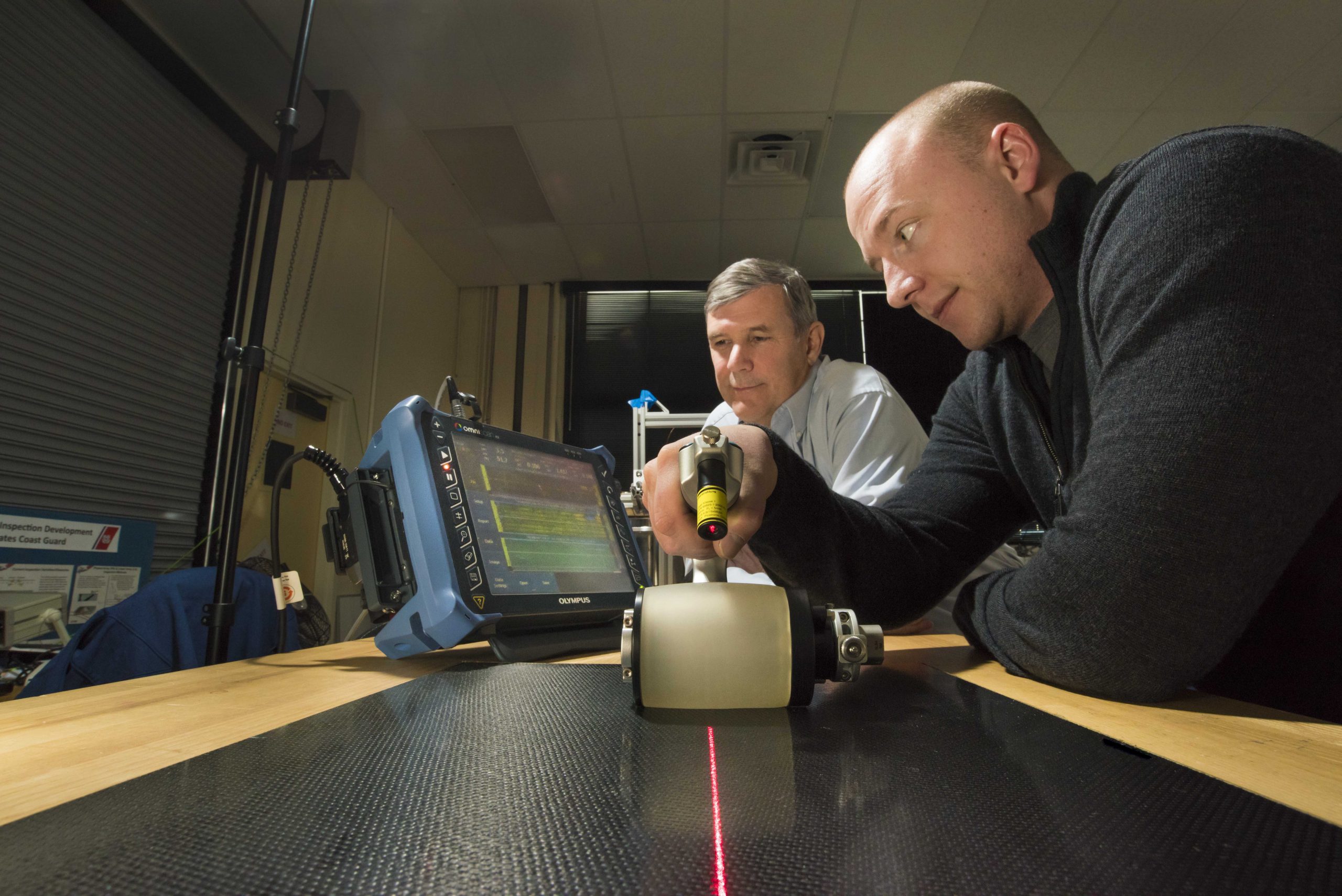 Sandia National Laboratories technologist Andrew Lentfer passes a roller probe over a composite as researcher David Moore checks data on a screen. The nondestructive testing technique sends sound waves into the composite material, returning data with each swipe of the roller probe.