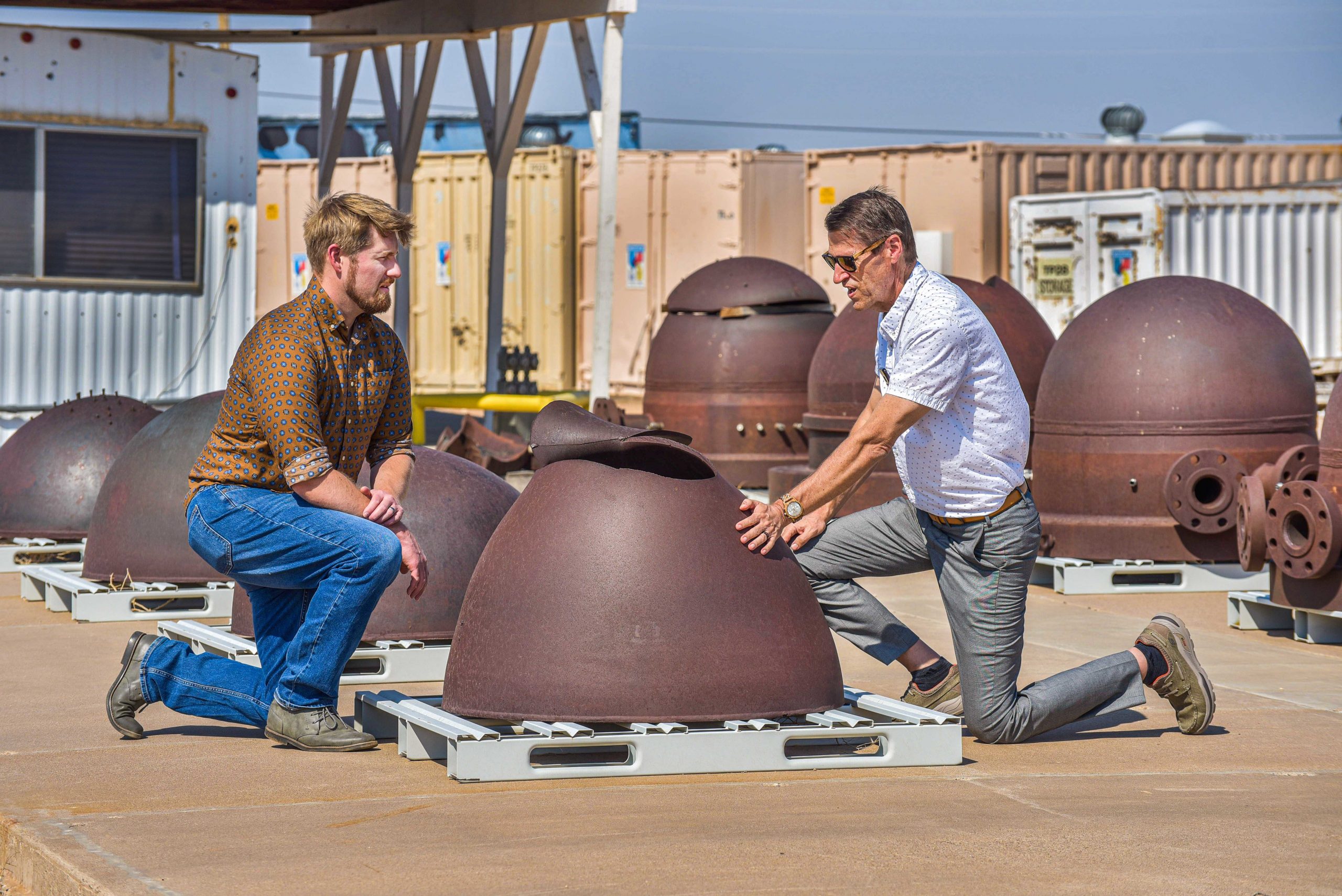 Two men, one younger and one older, look at a large, upside-down metal bowl with a huge rip in it.