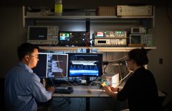 A man and a woman sit in a darkened room illuminated by computers and miscellaneous electronic equipment.