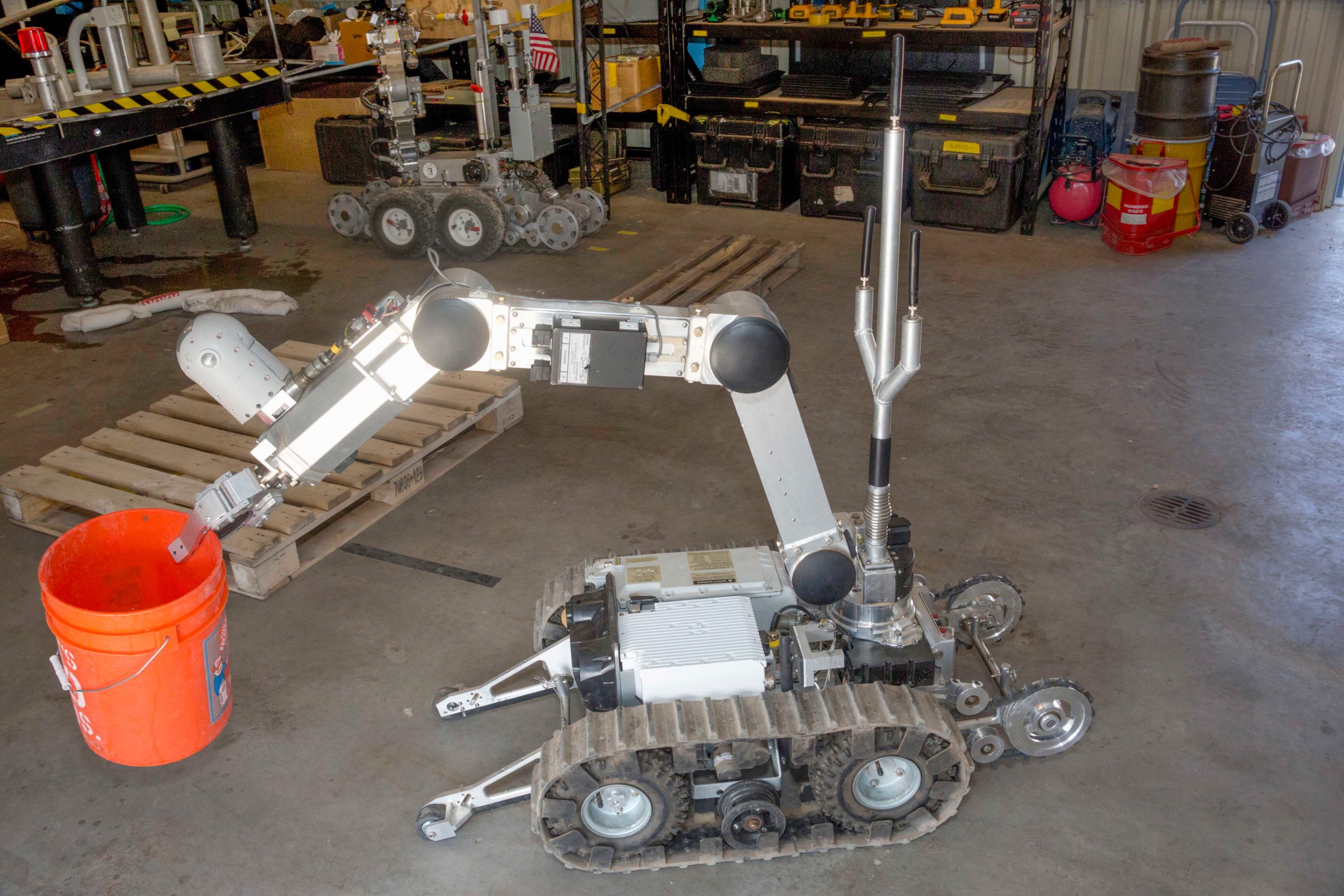 Foreground: Robot holding an orange bucket. Background: Puddle of water and a robot by some pipes. General messy workshop space around.