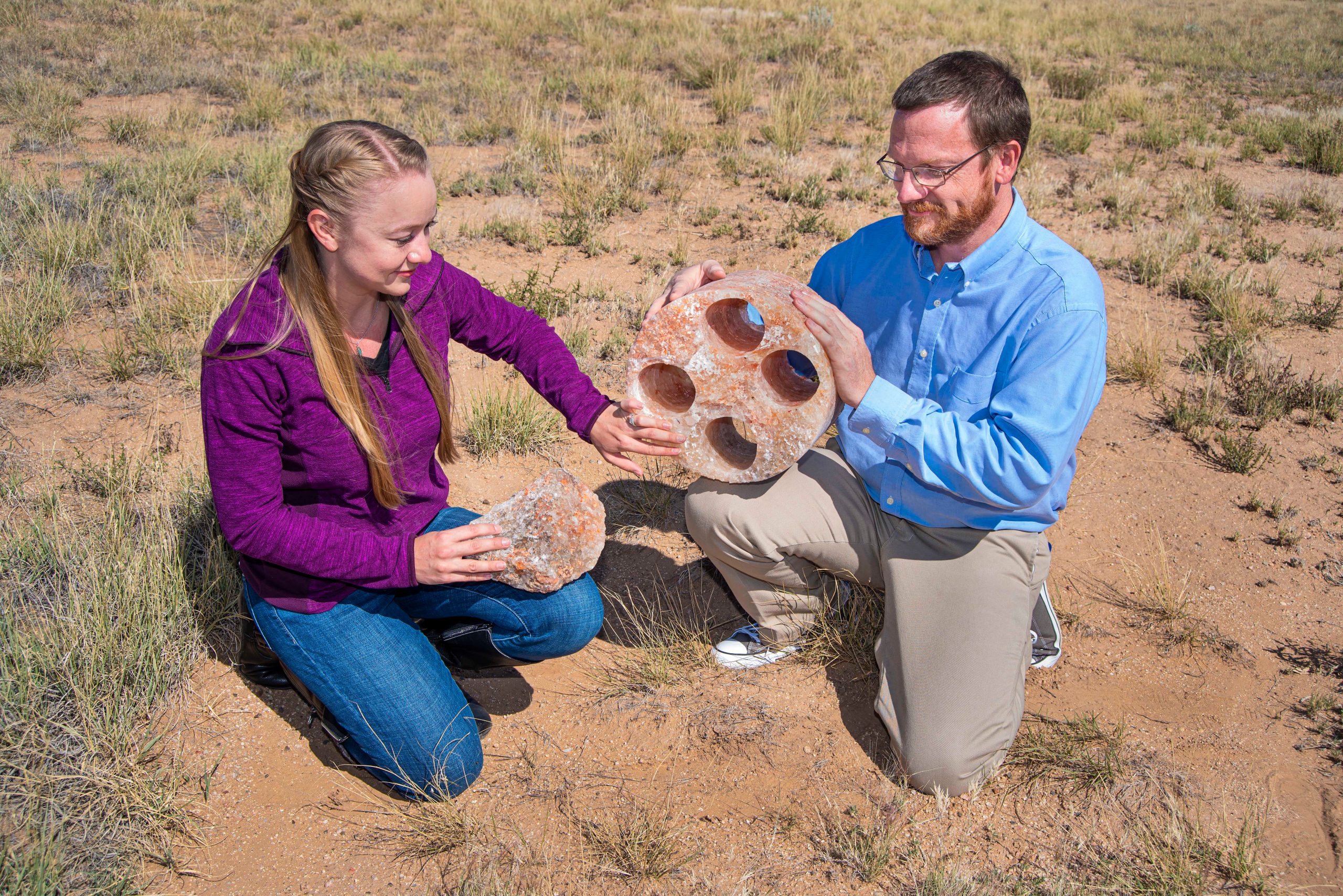 Two people holding large hunks of pinkish salt. One is a cylinder the size of a basketball. The other is more rough, the size of a softball.