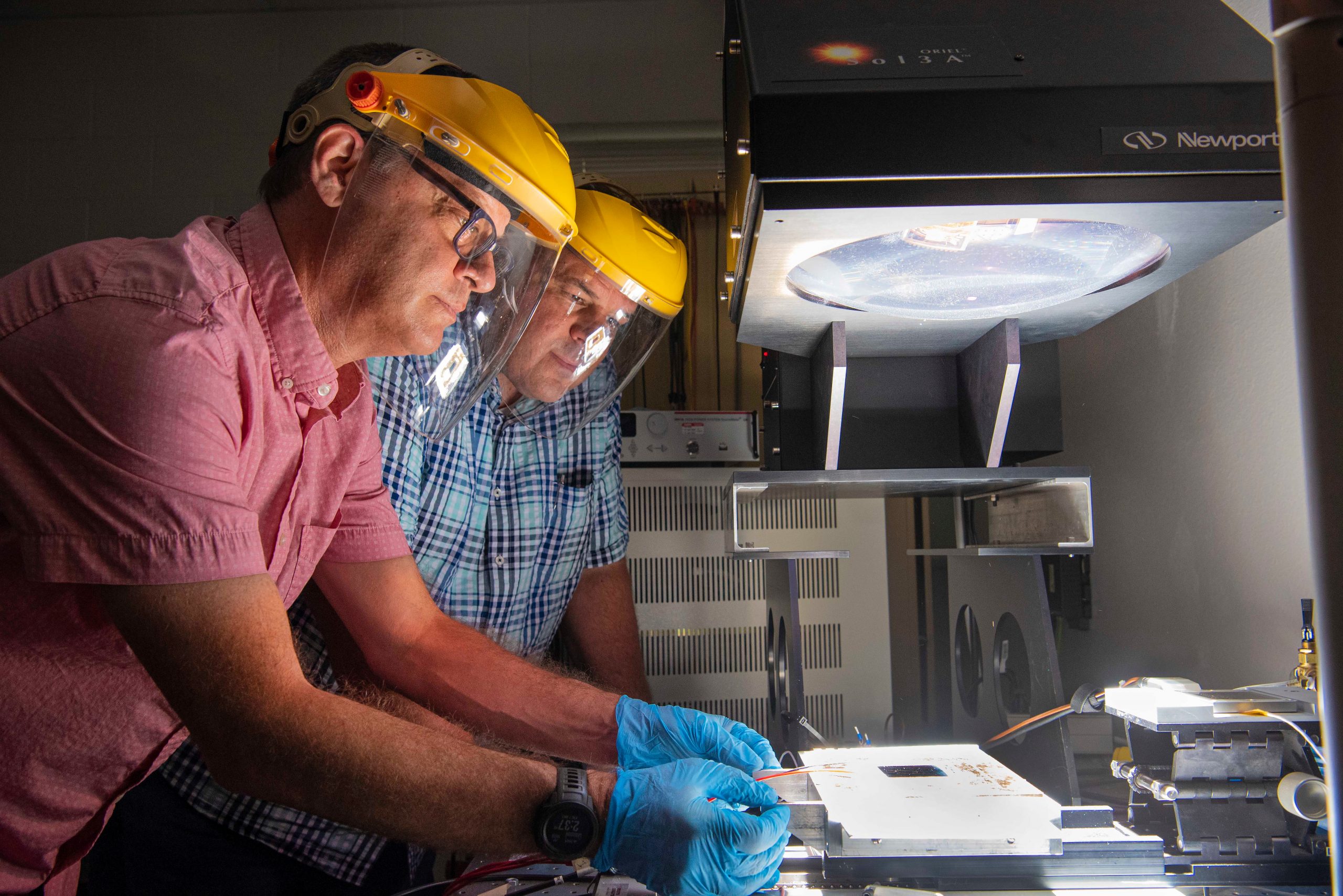 Two men in protective helmats look at a solar module under a bright light.