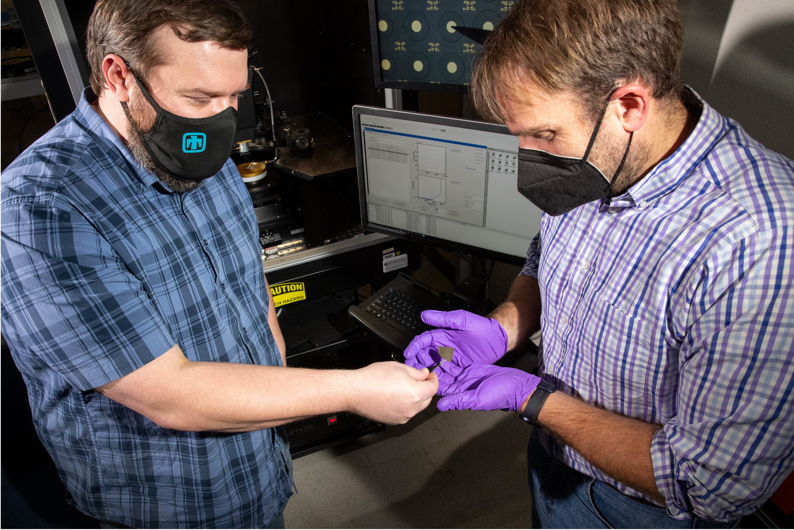 Foreground: Two men, one passing a nickel-sized wafer. Background: A device for testing the diodes on the wafer and a computer screen showing an array of dots/diodes