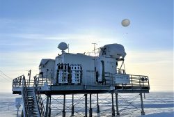 A metal building on stilts, with a white balloon taking off, above a snowy plain.