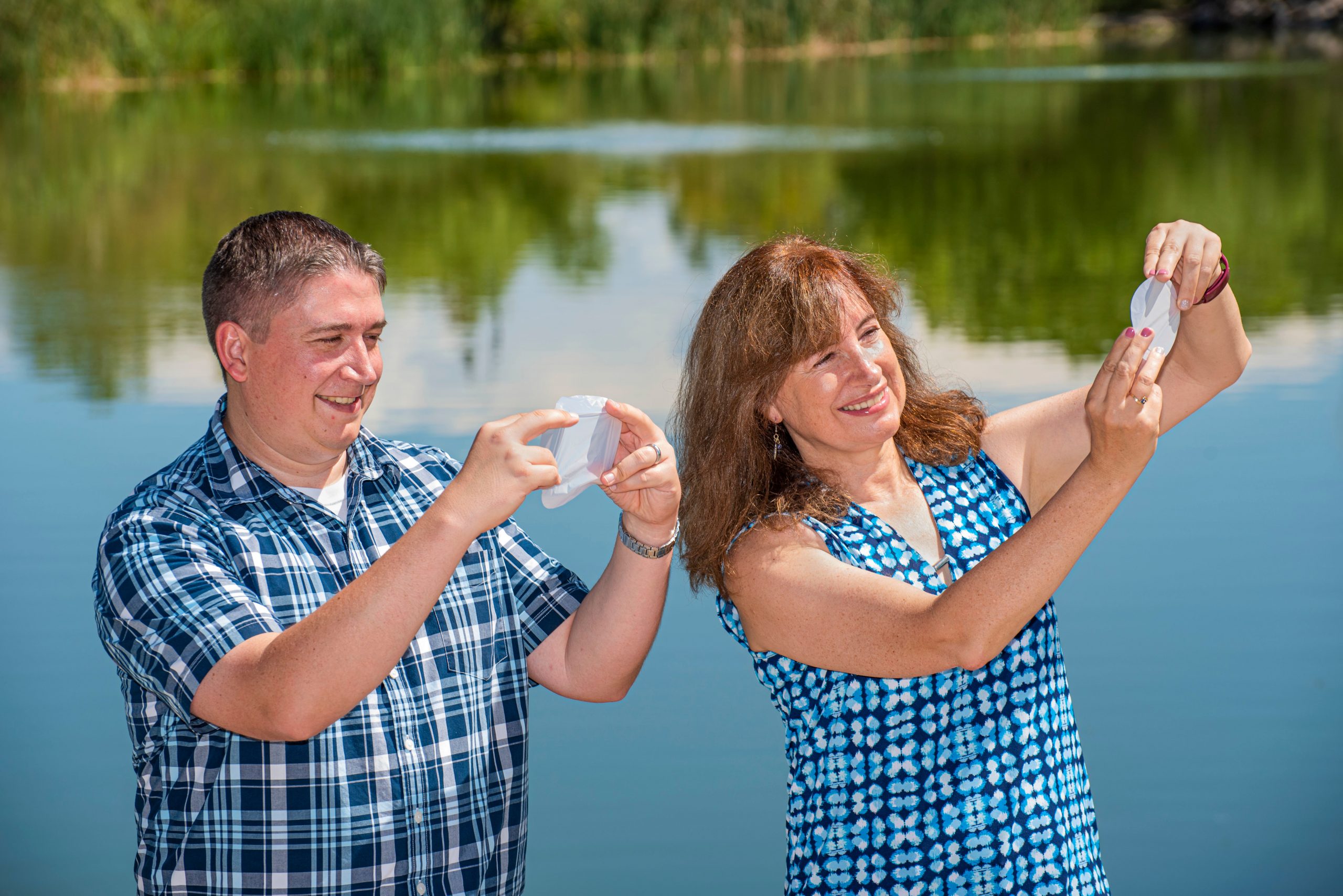 Two scientists look at hand-sized white membranes, water and lush trees in background.