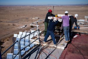 Four men in hard hats gathered around a rectangle. Far below mirrors can be seen.