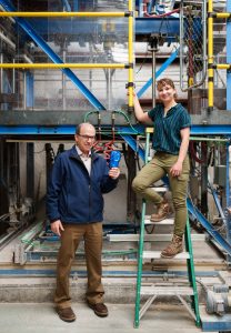 A man and a woman stand in front of a complex machine. The man is holding a blue drill bit. The woman is on a ladder.