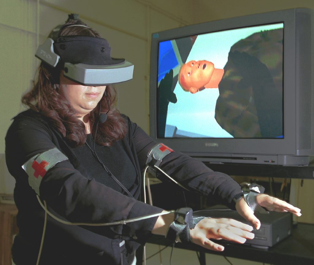 VIRTUAL LIFE-SAVER -- Lydia Tapia of Sandia National Laboratories demonstrates BioSimMER, a virtual reality application that allows rescue personnel to practice responding to a terrorist attack involving release of a biological agent. Through her goggles, Lydia sees the scene that is displayed on the screen behind her. The virtual patient she is treating suffers from a severe head wound and exhibits realistic symptoms. If she doesn't administer the proper medical treatment in time, the patient will die.