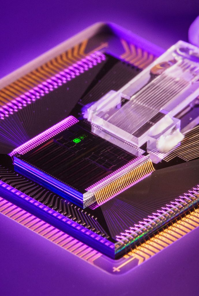 MINI MODULATOR — Sandia’s four-channel, silicon photonic single-sideband modulator chip, measuring 8 millimeters on each side and marked with a green Sandia thunderbird logo, sits inside packaging that incorporates optical fibers, wire bonds and ceramic pins. (Photo by Craig Fritz)